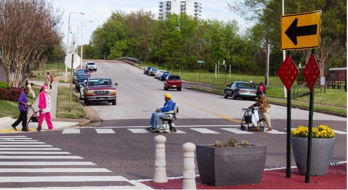 This image shows an intersection. From the angle shown, two crosswalks are visible. There is a person crossing in a motorized chair and another with a walker. Two other people are about to walk across the street. There are cars parked and trees in the background. In the foreground are two planters.