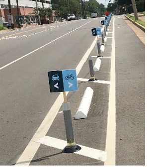 This image shows a street. There is a building on the far right, then a multi-lane road, then a parking area, then a separated bike lane, then a sidewalk. There are several signs in between the parking area and bike lane that show where cars and bikes should go, respectively.