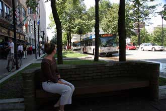 Photo of person sitting on bench on green area aside of road.