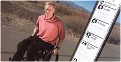 Photo of Peter Axelson in his wheelchair along a trail marked with sign detailing trail accessibility.
