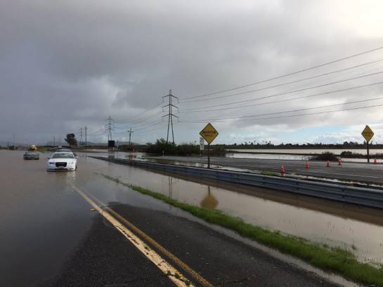 Cars drive on a flooded road next to a body of water.
