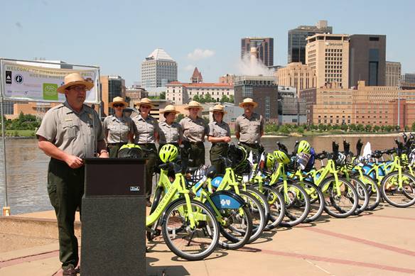 Park rangers stand next to bike share bikes next to the Mississippi River, with a city view in the background.