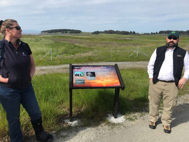 Two people stand on either side of an interpretive sign with marsh in the background.