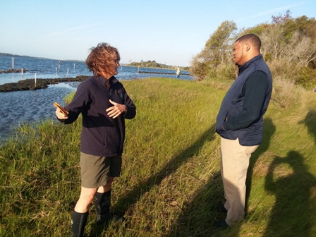 Two people talking and standing in marsh grasses.  Water and rock sill visibile in background.