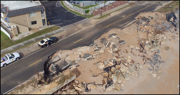 Shown from above, the highway broken along the edge near the beach where asphalt has fallen down the slope. One lane is unusable.