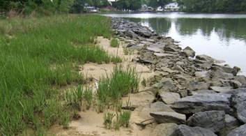 Photograph of a rocky sill creating a buffer between a body of water and sand and beach grass. The rocks fade into the sand on one wide and into the water on the other.