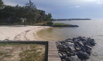 Photograph of the Perdido Bay pocket beach. Two rocky bulkheads stretch several meters into the water from a small sandy beach, their arms stretching toward each other to form a protective reach. A small line of timber is situated at the base of the bulkhead along the shoreline.