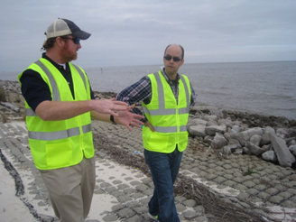 Photograph of two men in safety vests walking and talking along a rocky shoreline.