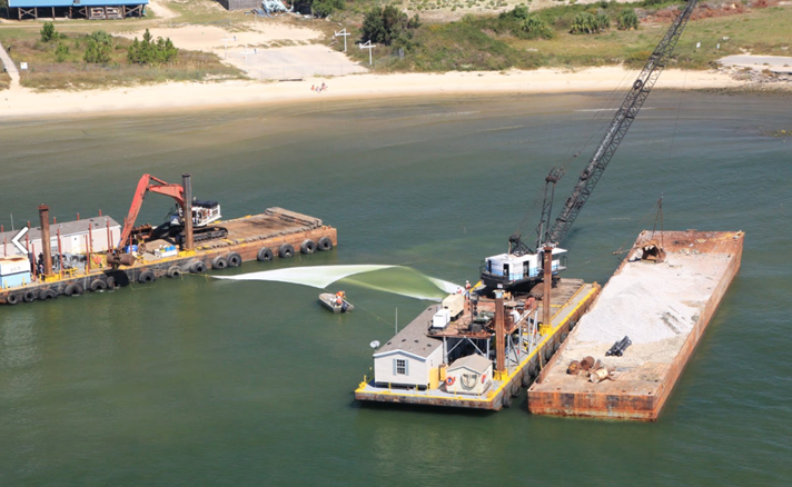 A photograph showing three construction barges staged offshore of a beach nourishment project site. The barges are being used to anchor opposite ends of a large sheet of geotextile fabric that is being deployed on the bottom to serve as a filter layer below the foundation of a breakwater structure.