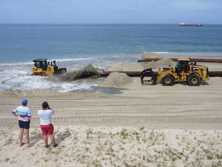 Photograph of an active beach nourishment project in the foreground and the Atlantic ocean and a hopper dredge ship in the background. On the beach are two dredge pipe outfalls with a mixture of sediment and water discharging from one direction onto the beach. A track-mounted bulldozer is shaping the discharged material.