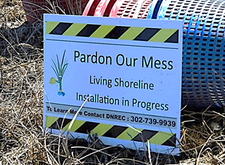 A picture of a sign at a living shoreline site that reads, "Pardon Our Mess: Living Shoreline Installation in Progress."