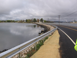 Photo shows roadway with water on either side and guardrail.