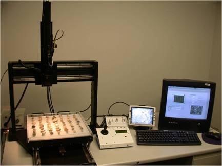 Figure 1.1. Photo. A picture of AIMS1. Research Aggregate Imaging System equipment (AIMS1). Left to right on a table, are a computer screen displaying text and graphics; a keyboard; a second smaller screen with display; a white, low device with a narrow display window and a joystick; and at the end, a white square tray containing pieces of aggregate arranged in seven even rows. The tray is positioned on a platform at the base of and within a large open upright framework–one upright member at either side and a larger, flat bar across the top on which a tall black metal device is mounted that appears able to move back and forth over the tray. This device is plugged into a small box behind the apparatus.