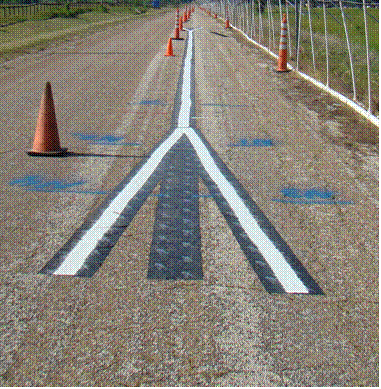 This picture shows a general southbound view of the rain tunnel with a test marking installed. A black polymer thin film is placed directly underneath the test markings to allow easy removal after the experimentation. The rain nozzles are on the right shoulder, and cones are placed to protect the test markings from any daily traffic. The test marking is longitudinal in the middle, and on both ends, it bifurcates to generate two possible lane shift directions. The closer end to the camera shows two extensions forming an upside down letter "V" shape,which then turnes into a longitudinal single solid lane, and at the other end, splits again to form a V shape.