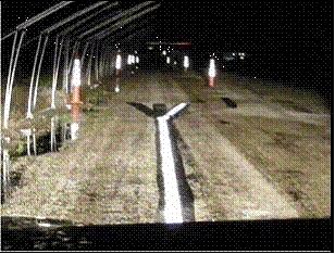 This picture shows a view of the pavement marking installation at night from inside the vehicle with low-beam headlamps. The cones are placed on both sides of the travel lane in a staggered formation. The piping for the nozzles are on the left shoulder. The test marking is longitudinally placed in the direction of travel directly underneath the vehicle, and at the end, it bifurcates into two extensions (or tapers). The left taper is covered with the masking tape, and the right taper is open and visible.