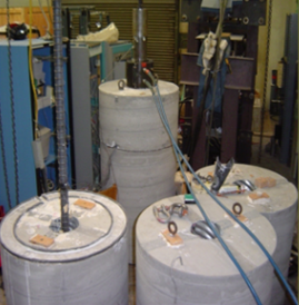 Figure 9. Photo. Test setup for large-bar anchorage testing. Four large bar anchorage test specimens show the test setup consisting of a cylindrical block of reinforced concrete with a corrugated metal pipe blockout. Reinforcing bars are then grouted into the blockouts and tested until failure using a hydraulic ram.