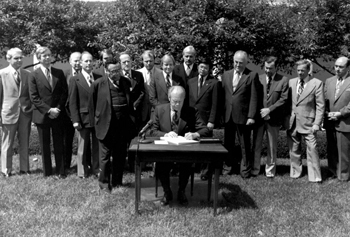 Signing Ceremony: Secretary of Transportation William T. Coleman, Jr., looks on as President Gerald R. Ford signs the Federal-Aid Highway Act of 1976. It established the Interstate 3R Program (resurfacing, restoring, and rehabilitating), creating a 'Transition Quarter' as the Federal Government shifts the start of the fiscal year from July 1 to October 1, revising the Interstate withdrawal/substitution provision of the 1973 Act to allow substitute highway as well as public transportation projects. Immediately behind the President are Representative Norman Y. Mineta (CA), a future Secretary of Transportation (over the President's left shoulder), and Federal Highway Administrator Norbert T. Tiemann (behind Mineta).