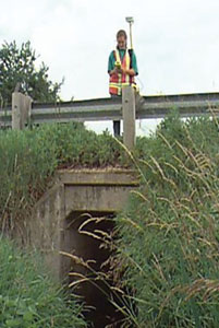 Photograph. A field inspector uses a global positioning system receiver to collect data that will be recorded in HYDINFRA. The inspector is standing on the roadway above a concrete culvert.