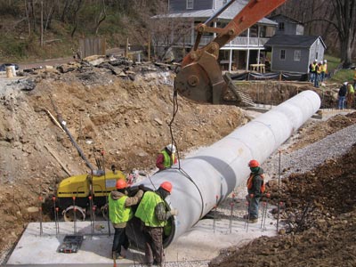 Photograph. Workers are installing a long section of round pipe that appears to be between 4 and 5 ft across and made of concrete.