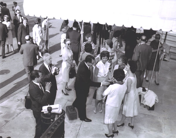 Mrs. Rex Whitton, wife of the Federal Highway Administrator, looks on (in sunglasses) as Deputy Federal Highway Administrator Laurence Jones greets Cabinet wives before the Landscape-Landmark Tour.  Next to him is Mrs. Mary Connor (facing camera, wearing necklace with disc to waist), wife of Secretary of Commerce John T. Connor. 