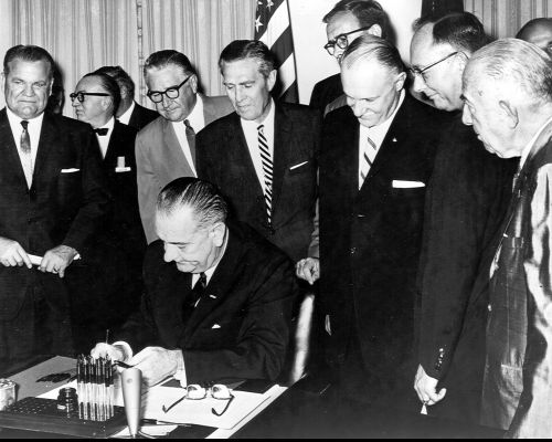 On August 13, 1964, Administrator Rex Whitton (in front of the flag) watches as President Lyndon B. Johnson signs the Federal-Aid Highway Act of 1964.
