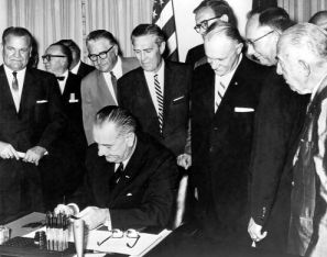 On August 13, 1964, Administrator Rex Whitton (in front of the flag) watches as President Lyndon B. Johnson signs the Federal-Aid Highway Act of 1964.