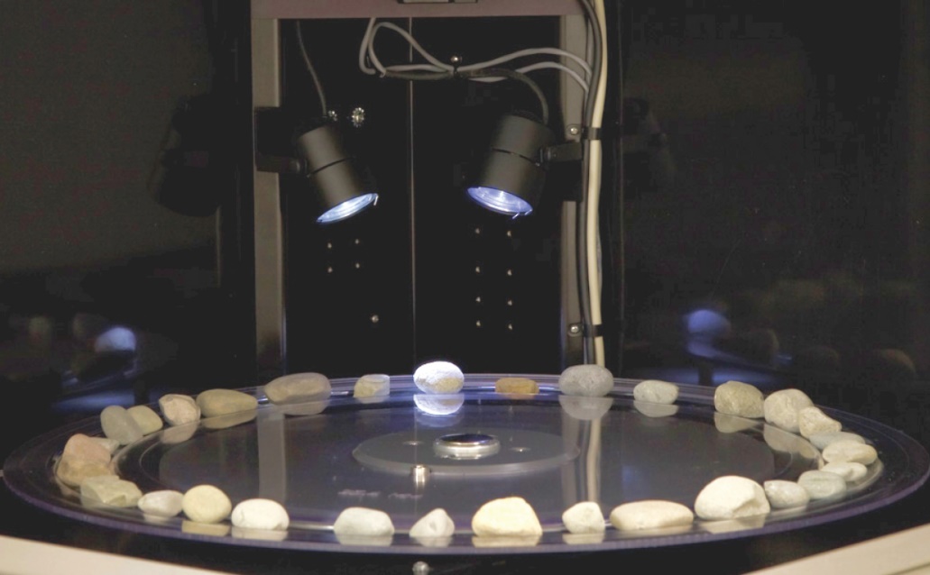 The photo depicts the inside view of the Aggregate Image Measurement System (AIMS2), an integrated hardware/software system that automates the process of measuring the particle angularity, form, and surface texture. Multiple aggregates are placed on a tray and the system captures and analyzes each material sample.