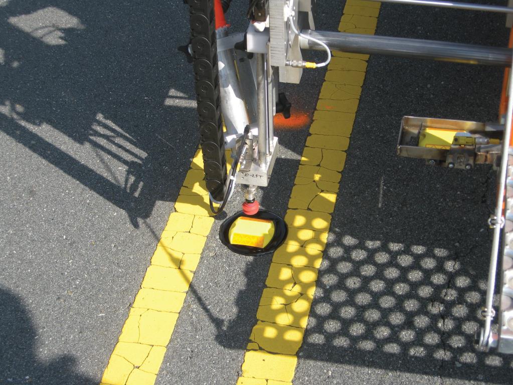 The photo depicts the installation of a pavement marker. As the applicator vehicle nears the point of installation, the operator engages the system and as the suction cup lifts the marker into place, the bituminous is placed onto the surface seconds before the reflective payment marker is placed. Once installed it returns back to the nest.