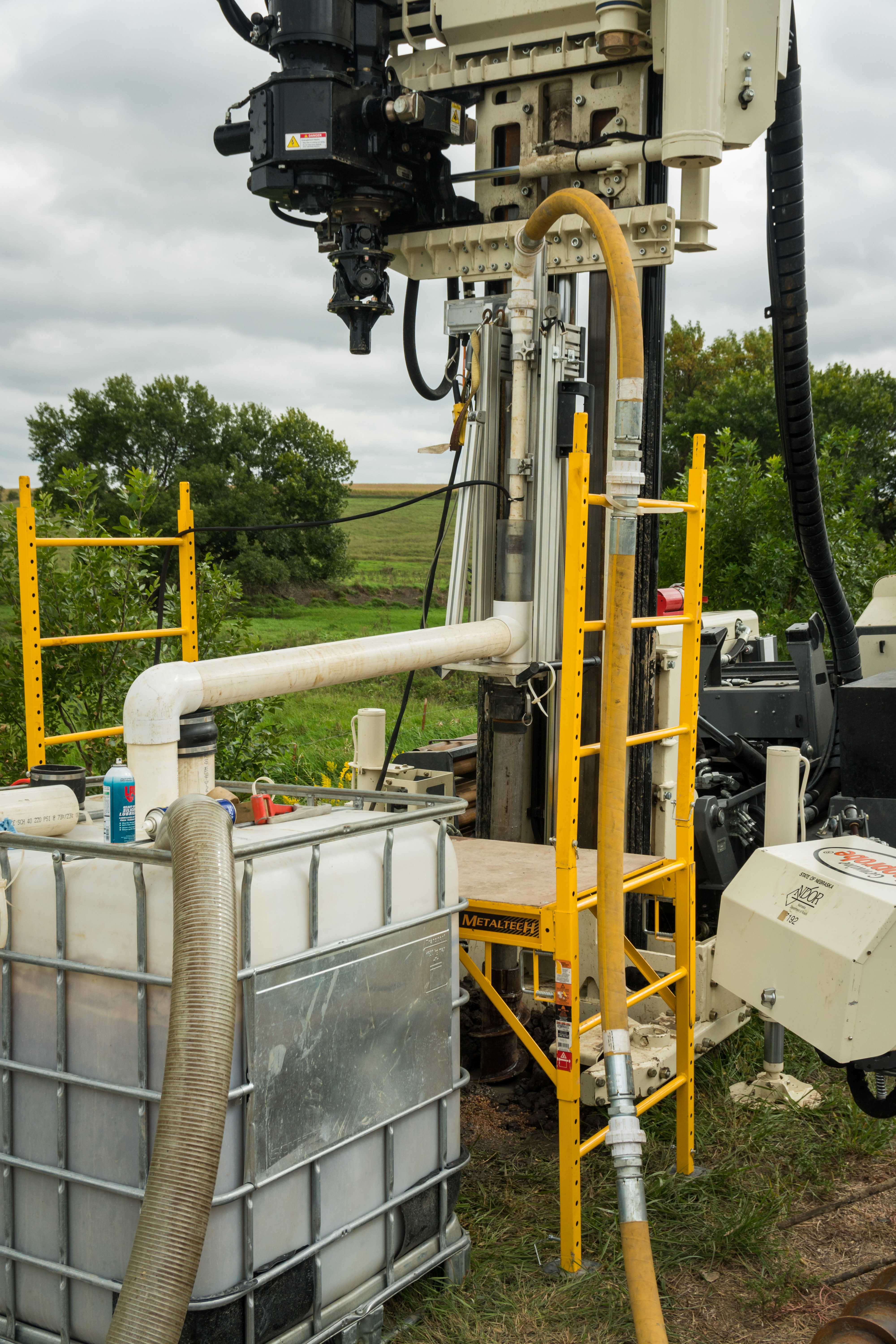 The photo depicts the typical In-Situ Scour Testing Device (ISTD) test setup in the field. The Advanced Linear Drive is shown mounted on top of the casings, which protrude from the auger section in the ground. The drill rig, used to auger the borehole for the test, is located behind the ISTD equipment. A series of flexible hoses attaches to the top of the device, where they connect to a 90-degree bend and then to the PVC piping mounted to the linear drive. The outlet PVC piping runs from the Advanced Linear Drive to a water tank shown in the foreground. A suction hose runs from the water tank back to the pump. A scaffold is assembled between the drill rig and the water tank to assist the technicians in setting up the device.