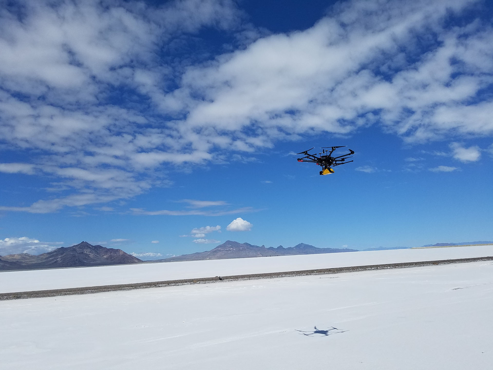 Image of a drone leaving the ground with blue sky, clouds, and mountains in the background.