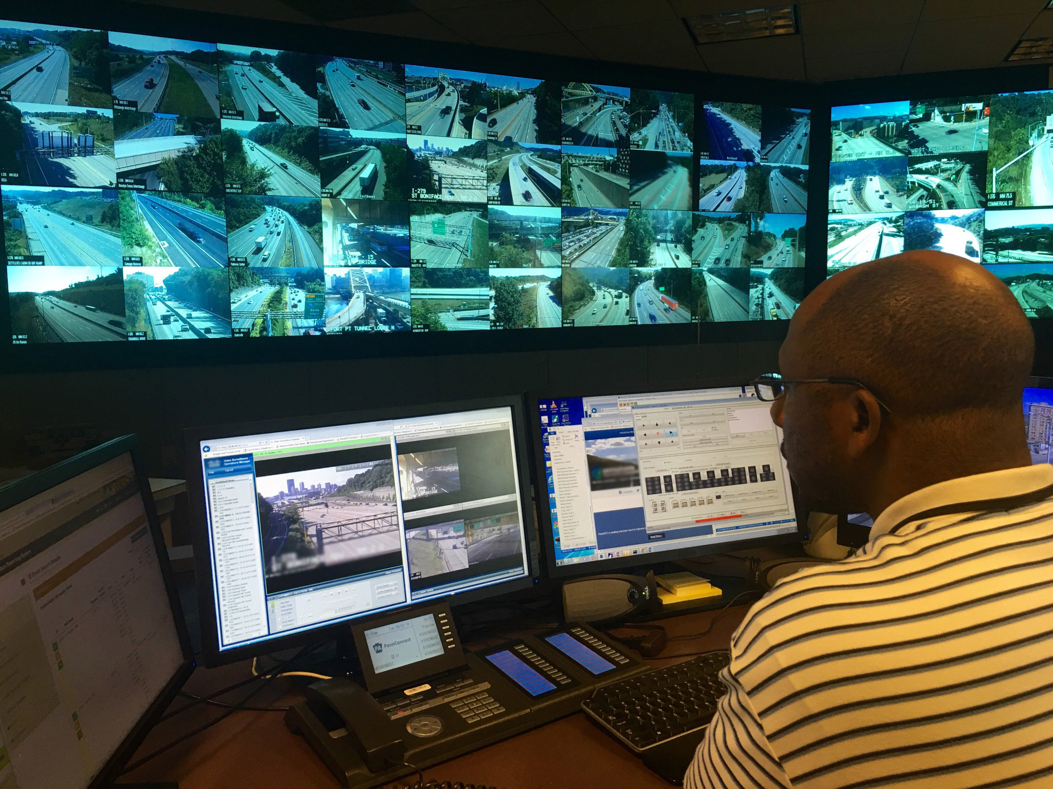 Image of a man sitting in front of two computer monitors with a wall of traffic monitors in the background.