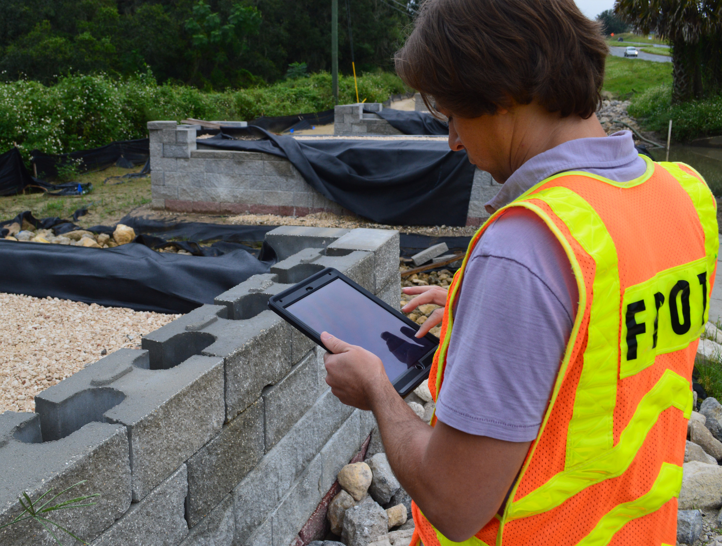 A man in a safety vest standing in front of a cement block wall and working on a tablet.