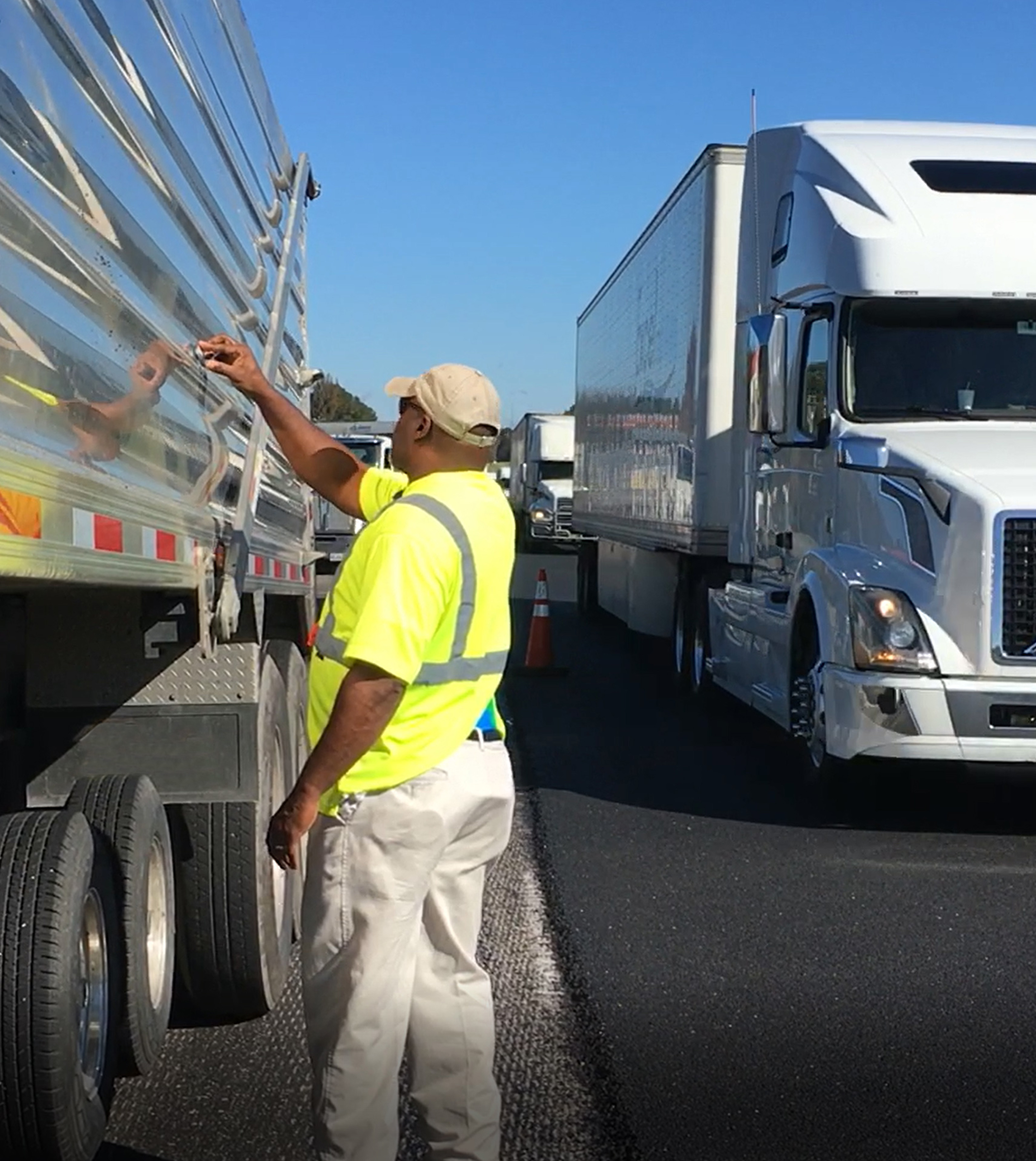 A man on a roadside standing next to a dump truck with several semi trucks approaching on the road next to him.