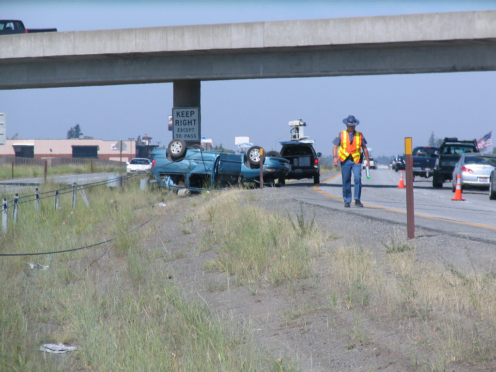 A state trooper in a safety vest walking along the side of a highway at the scene of a crash where a car is flipped upside down.