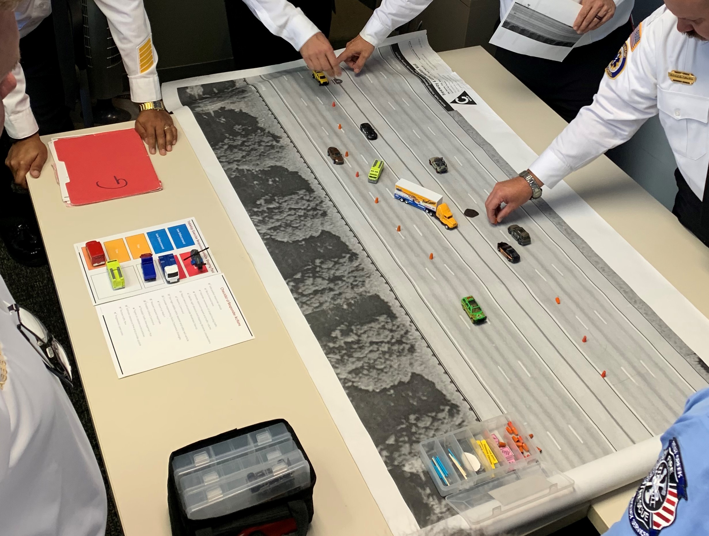 Multiple people standing at a tabletop learning exercise with a large printout of a roadway and toy vehicles to practice traffic incident management scenarios.