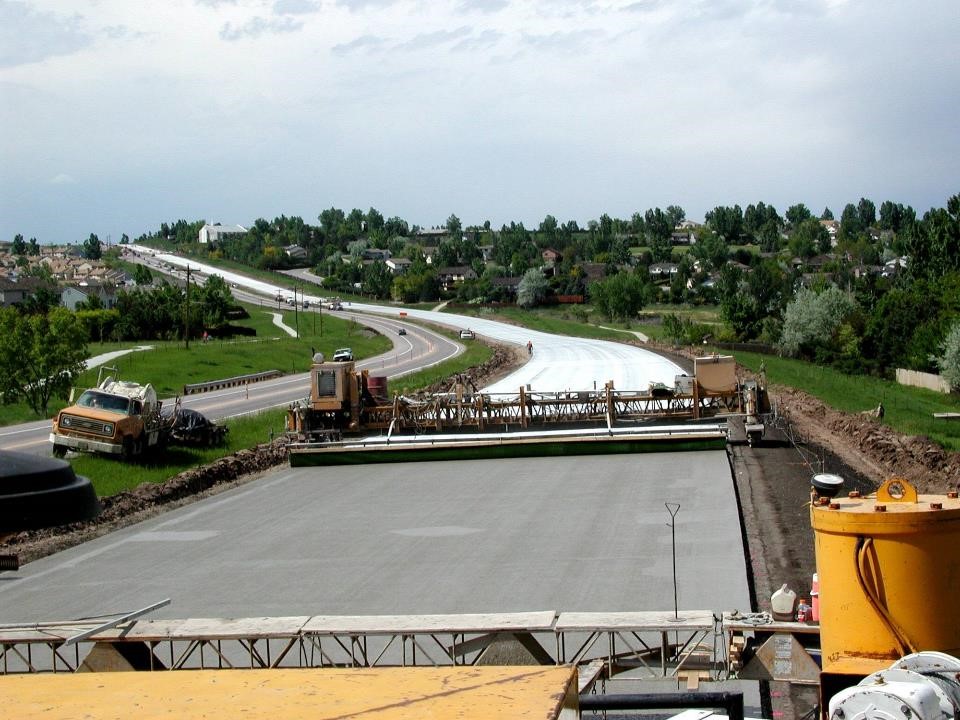 Two lanes of a four-lane highway are under construction. Concrete paving equipment can be seen in the foreground. Houses are shown on either side of the highway. 
