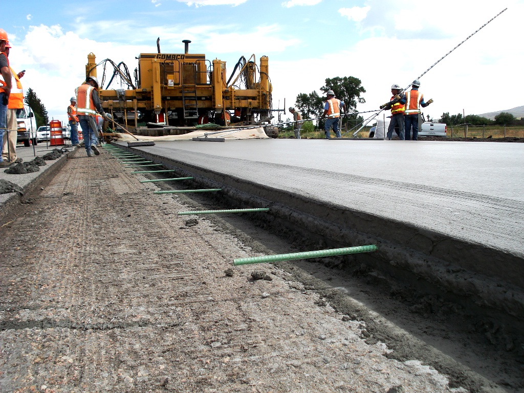 Ground-level view of crews brushing newly formed concrete pavement in the distance. Eight workers are shown behind paving equipment.