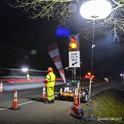 A photo of a highway worker in high-visibility yellow gear standing near a portable traffic signal with an arm to stop traffic in a nighttime highway work zone. The signal displays a two-signal traffic light above a 'STOP HERE ON RED' sign. Long-exposure light trails are present from passing cars and the traffic arm in motion under the street light.