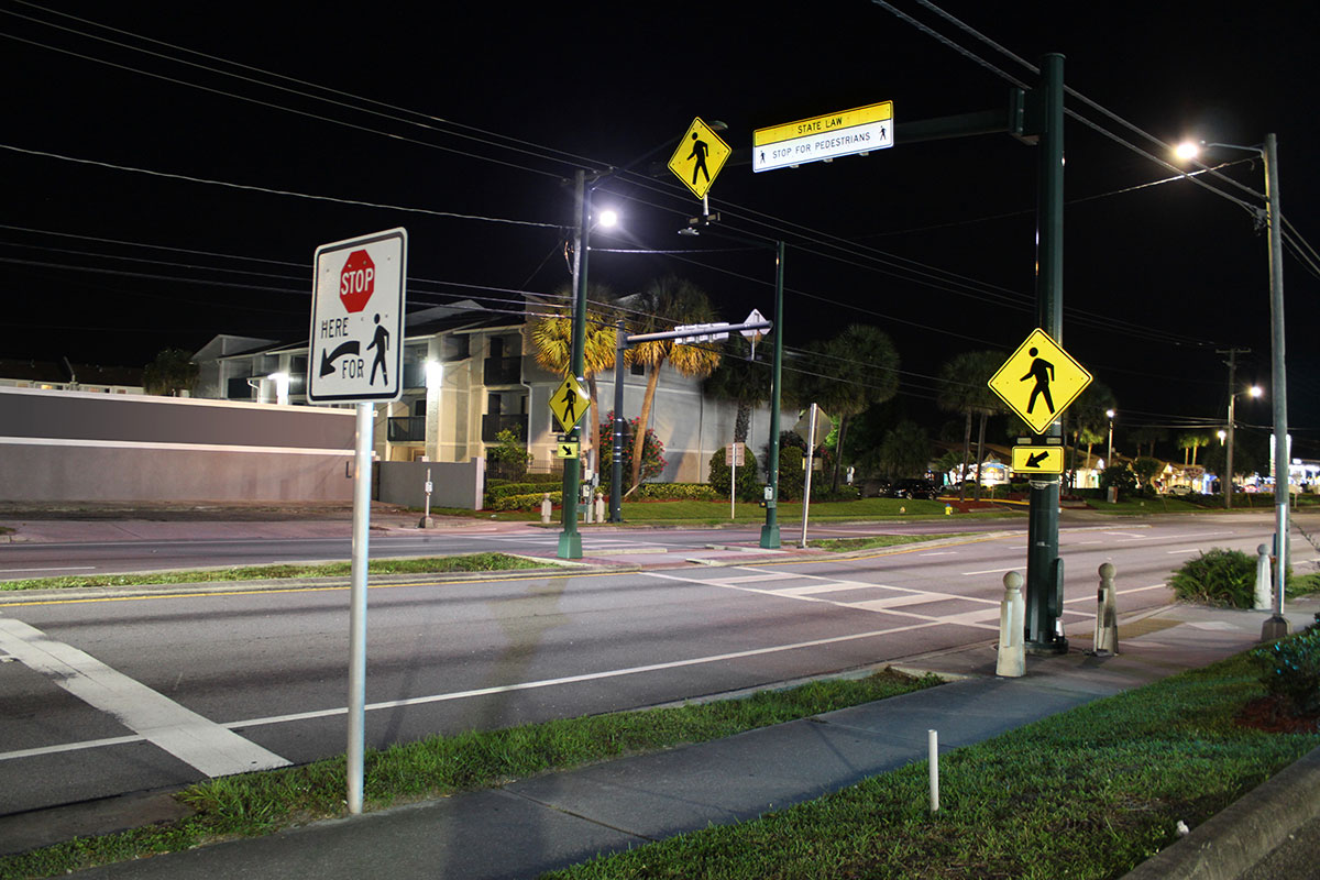 Roadway crosses left to right. A crosswalk goes across the road. Crosswalk is brightly lit and features multiple signs to alert motorists of its position.