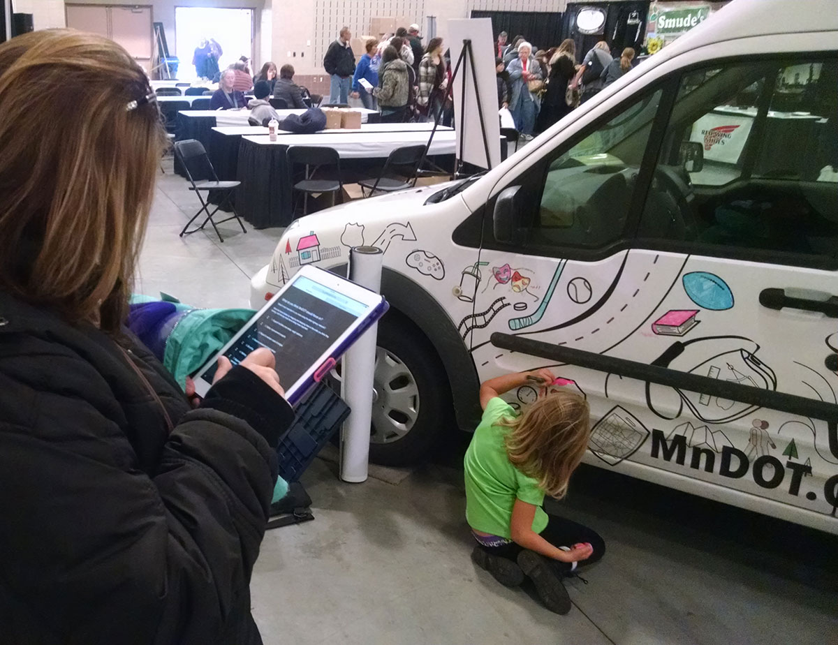 A child colors on a car featuring transportation planning themes while an adult completes a survey on a tablet nearby at a MnDOT pop-up event
