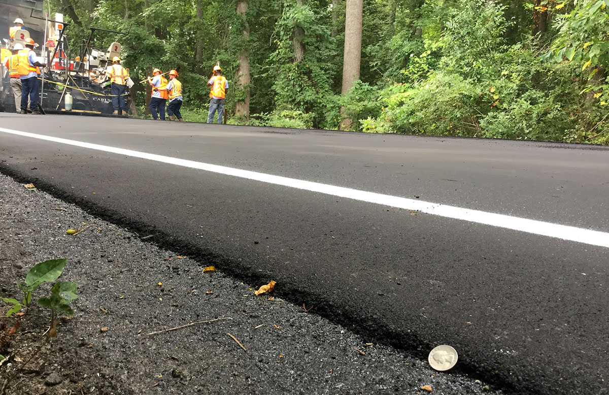 Thin asphalt overlay with quarter standing on its end showing depth of overlay. Installation crew is visible in background.