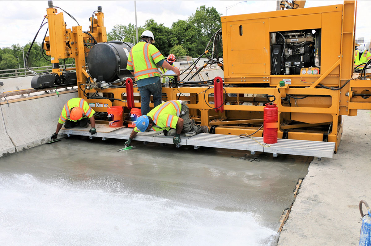 Placement of UHPC on a bridge deck. A UHPC paver machine is shown with workers finishing the UHPC.