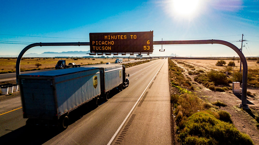 Truck driving on highway passes under dynamic message sign indicating minutes to: Picacho 6, and Tucson 53.