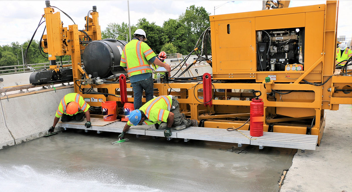 Placement of UHPC on a bridge deck. A UHPC paver machine is shown with workers finishing the UHPC.