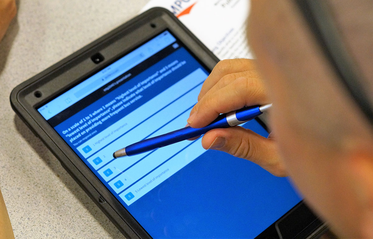 A hand holding a stylus makes selections on a tablet depicting a digital survey.