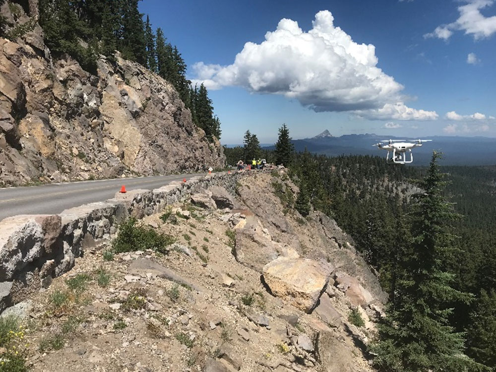 An aerial drone hovers near a cliffside highway near workers wearing high visability vests.