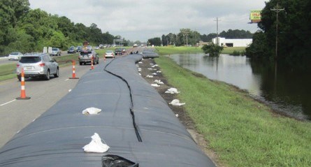 At right, flooded low-lying area. A water-inflated barrier extends through the center of the image, at the roadway edge. At left, vehicles use the roadway, protected by the inflatable barriers.