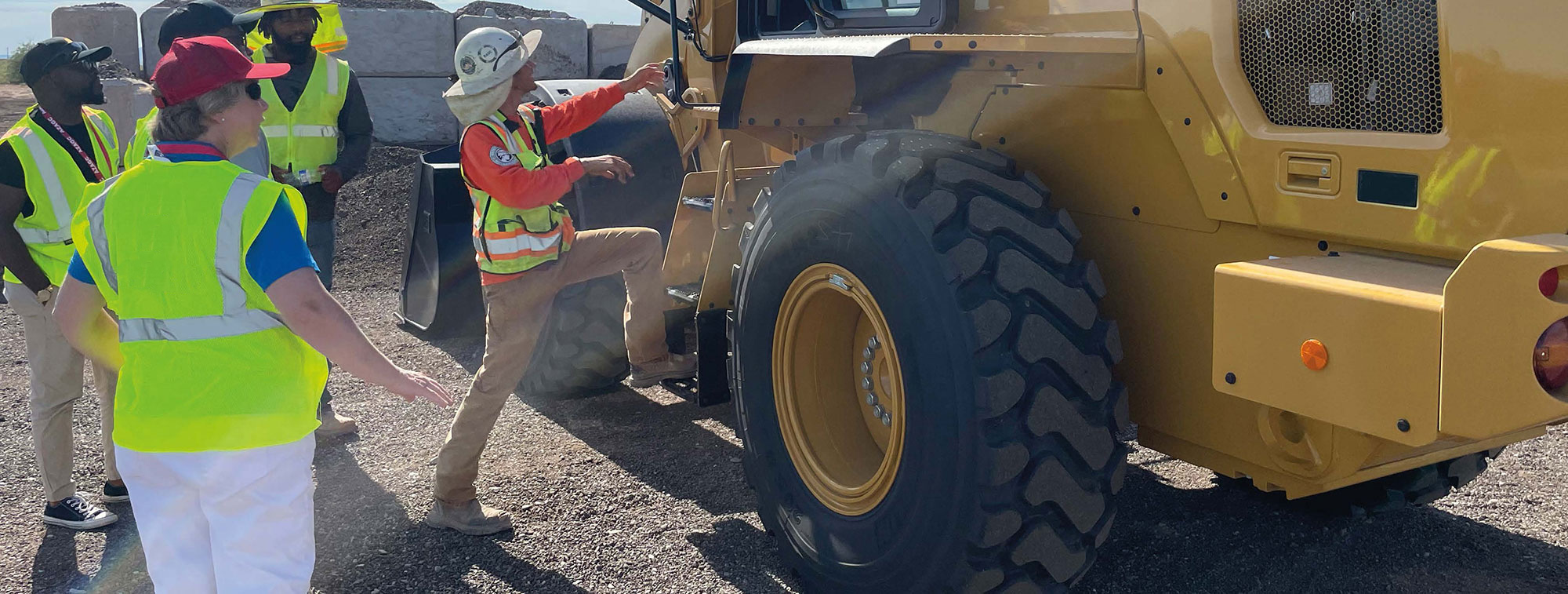 At left, four people, wearing safety vests, look on as another person, at right, wearing safety vest and hard hat, climbs into a piece of heavy equipment.