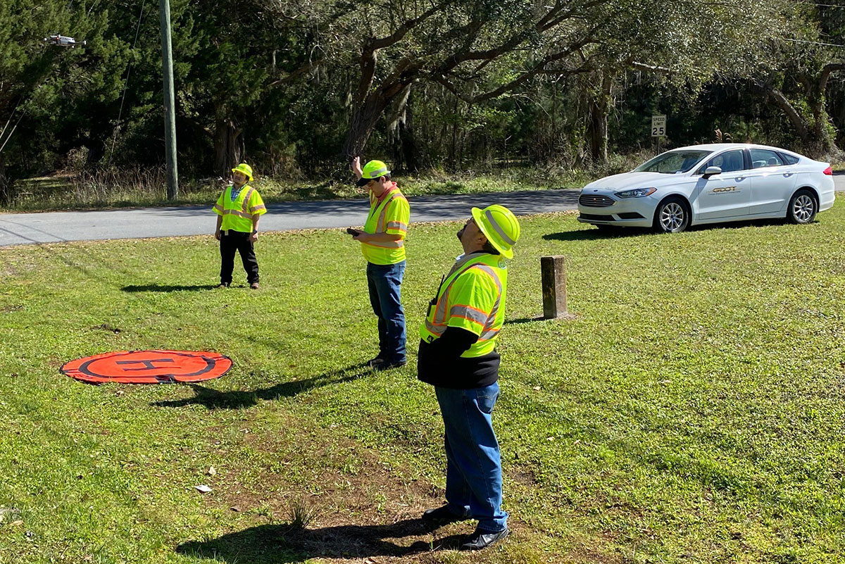 3 men standing in a grassy area around an orange UAS landing pad in hard hats and reflective vests. A UAS hovers at top left.