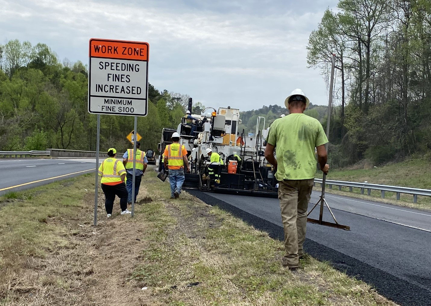 4-lane divided highway with work crew installing asphalt on the inside lane of the right side of the image. An inspector stands in the median near a sign.